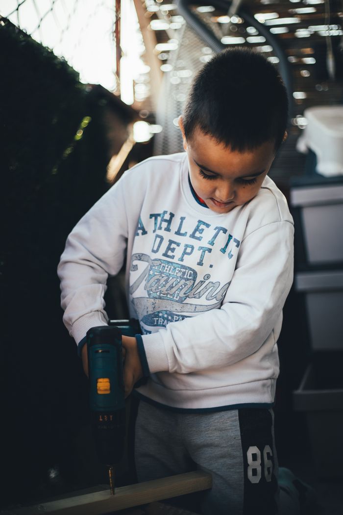 A young boy focuses intently on using a drill in a woodworking project indoors.