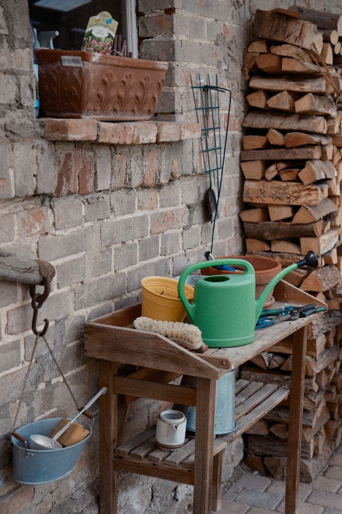 Rustic outdoor scene with gardening tools and stacked firewood against a brick wall.