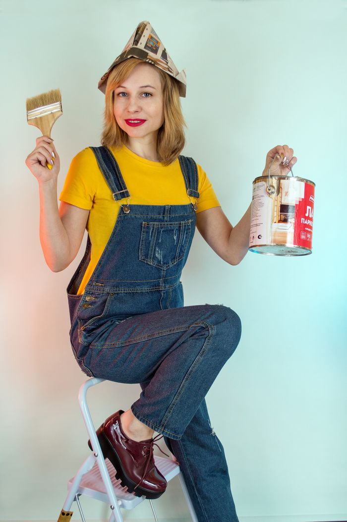 Cheerful female painter in denim workwear and paper hat showing brush and paint can while sitting on ladder and looking at camera